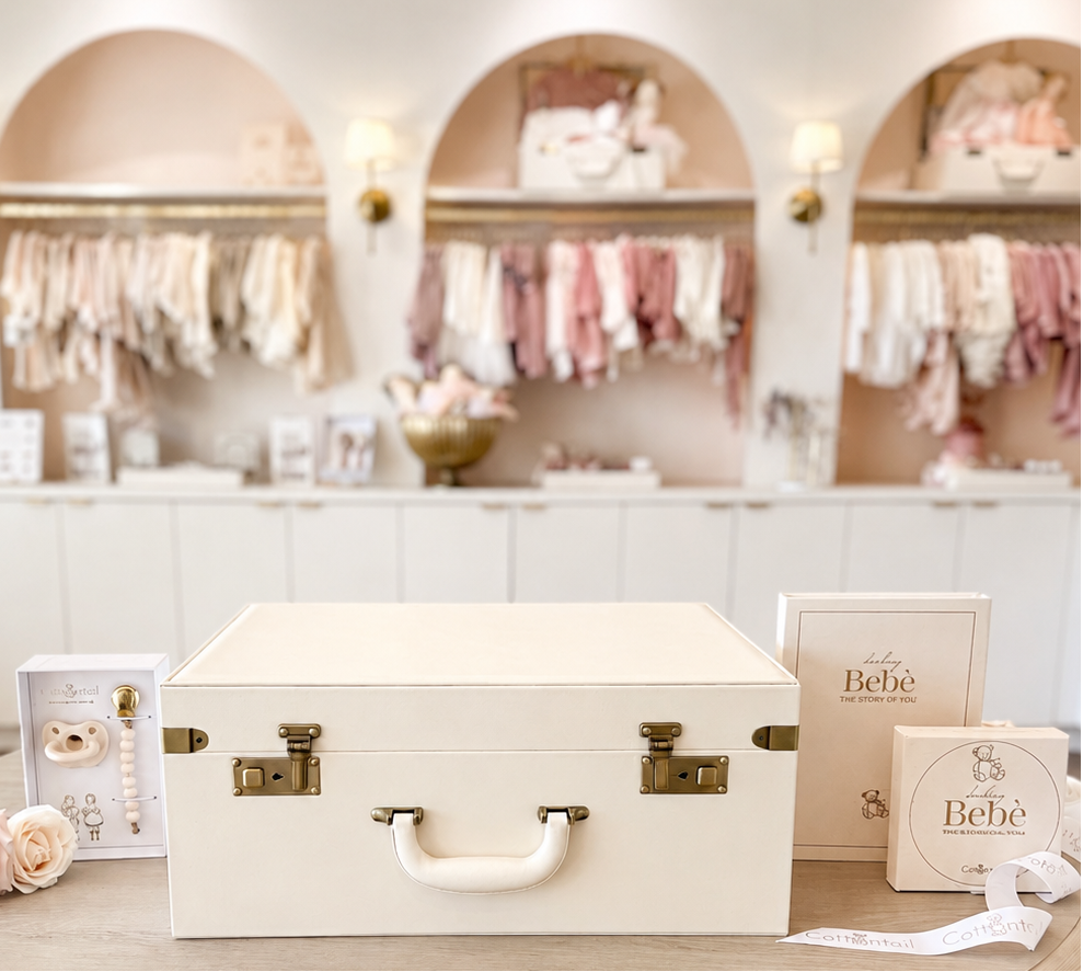 A cream-colored wooden storage trunk with brass latches and a white handle sits on a round wooden table in a boutique setting. The trunk is flanked by branded BeBe products, including a box and a small jar, with a pink rose decoration on the left.